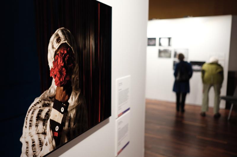 Blick in die Ausstellung ohne Personen mit einem der Siegerbilder aus dem Jahr 2024, auf dem sich eine Frau rote Blumen vors Gesicht hält, damit sie nicht erkennbar ist