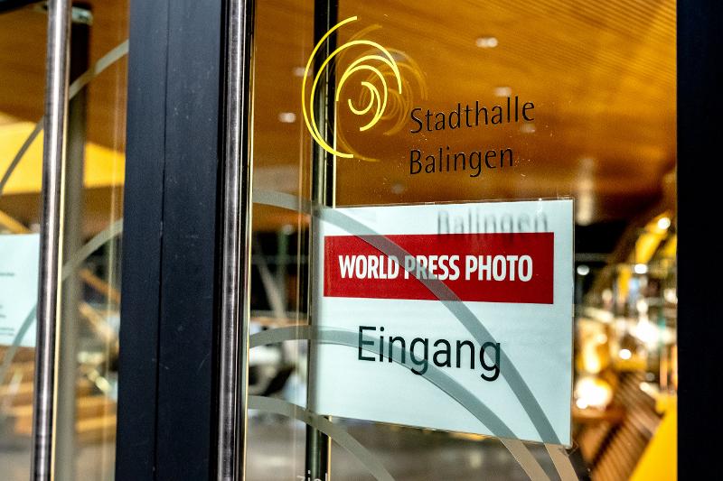 Schild mit dem Schriftzug "World Press Photo" Eingang an der Glas-eingangstüre der Stadthalle, im Hintergrund erkennt man unscharf das inner der Halle mit der markanten, schrägen Holzwand